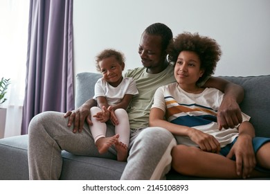 Black family watching TV or movie on sofa at home. Dad hugging daughter and son. Family relationship and spending time together. Fatherhood and parenting. Domestic entertainment and leisure. Daytime