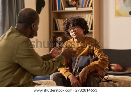 Black father gesturing while talking with teenage son wearing glasses and holding backpack at home, discussing school or family issues. Useful for parenting, counseling, teen support campaigns