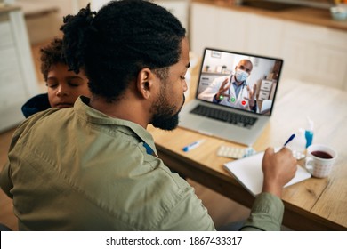 Black Father Having Video Call With Pediatrician And Writing Notes While Taking Care Of His Sick Son At Home. 