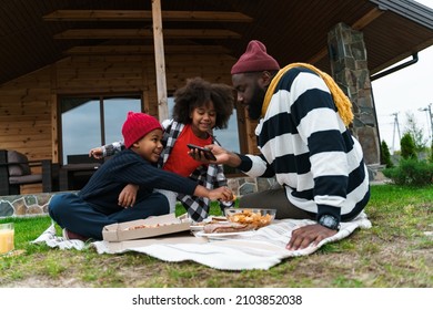 Black Father And His Sons Using Cellphone And Eating Chips While Resting Outside City