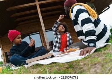 Black Father And His Sons Using Cellphone And Eating Chips While Resting Outside City