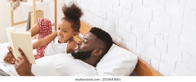 Black father and his young daughter are lying on a bed as they share a joyful storytime. The child listens intently, showing enthusiasm, while the father reads from a book, copy space - Powered by Shutterstock