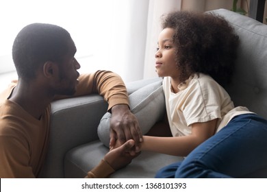 Black father hold daughter hand looking at kid girl lying on sofa feels unwell unhealthy upset, show express sympathy empathy, try to help with advice. Support, caring for sick child and love concept