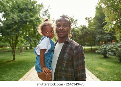 Black Father Holding And Hugging His Little Daughter In Arms And Looking At Camera On Pavement Road In Green Park. Happy Family Relationship And Spending Time Together. Fatherhood And Parenting