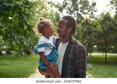 Black Father Holding And Hugging His Daughter In Arms In Blurred Green Park. Smiling Adult Man Looking At Little Girl. Family Relationship And Enjoying Time Together. Fatherhood And Parenting