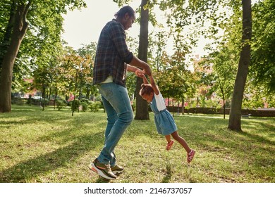 Black Father Holding And Spinning In Hands Hisjoyful Little Daughter On Lawn In Green City Park. Family Relationship And Spending Time Together. Fatherhood And Parenting. Summer Sunny Day