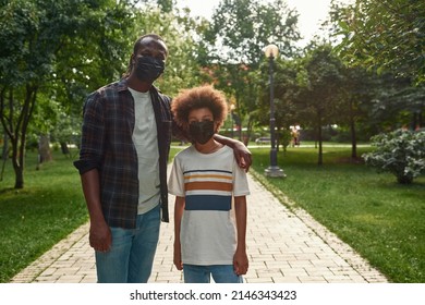 Black Father Hug Teenage Son On Pavement Road In Green Park. Man And Boy Wear Medical Masks Look At Camera. Family Relationship. Fatherhood And Parenting. Health Protection During Coronavirus Pandemic