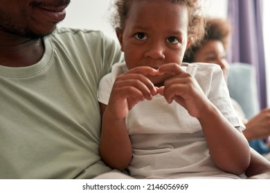 Black Father Hugging And Resting With Daughter On Sofa At Near Blurred Son At Home. Focus On Little Girl Showing Heart Gesture And Looking At Camera. Family Relationship. Fatherhood And Parenting