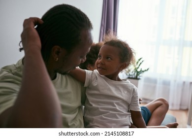 Black Father Hugging And Resting With Pleased Daughter On Sofa At Near Son At Home. Family Relationship And Spending Time Together. Fatherhood And Parenting. Domestic Lifestyle. Sunny Daytime