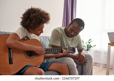 Black father looking at his teenage son playing on acoustic guitar on sofa at home. Family relationship and spending time together. Fatherhood and parenting. Domestic entertainment, hobby and leisure - Powered by Shutterstock