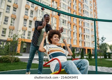 Black Father Swinging His Teenage Son On Swing On Playground Outdoors. Family Relationship And Enjoying Time Together. Fatherhood And Parenting. Entertainment And Leisure. Yard In Modern City District