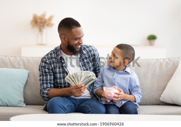 Black Father Teaching His Son Budget Planning And\
Financial Literacy Putting Money Savings In Piggybank Sitting On\
Couch Indoors. Personal Finances Concept. African Dad Talking With\
Kid About Money