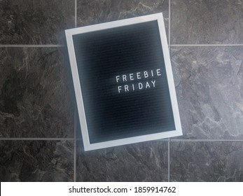 A black felt letter board with white frame is shot flat lay style on gray slate looking tile. Letters on the board are left justified and say Freebie Friday, a phrase businesses use for giveaways. 