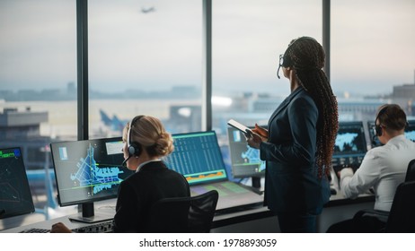Black Female Air Traffic Controller Holding Tablet in Airport Tower. Office Room is Full of Desktop Computer Displays with Navigation Screens, Airplane Departure and Arrival Data for the Team. - Shutterstock ID 1978893059