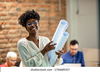 Black Female Architect Smiling In Busy Office. Female Architect With Blueprints. Portrait Of Confident Young Businesswoman Holding Blueprint In Office. Young Female Entrepreneur