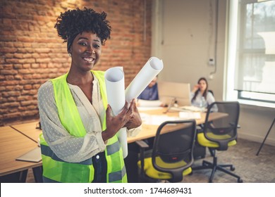 Black female architect smiling in busy office. Female Architect with Blueprints. Portrait of confident young businesswoman holding blueprint in office. Young female entrepreneur
