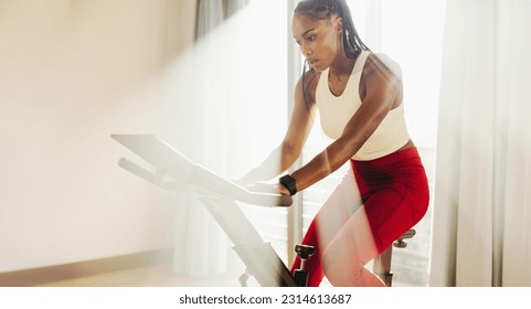 Black female athlete crushing her cardio workout with her home exercise equipment, using her drive and determination to achieve her fitness goals. Woman using a stationary bike for indoor cycling. - Powered by Shutterstock