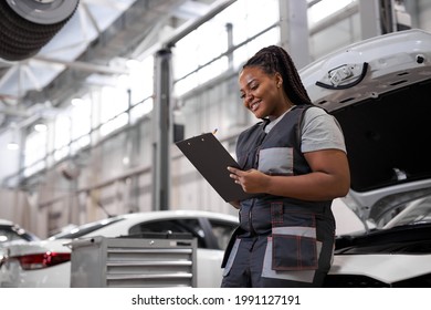 Black Female Auto Mechanic Making Car Checkup, In Uniform Writing And Holding Clipboard Of Service Order Indoors. Repair Service. Woman Car Repairing Concept