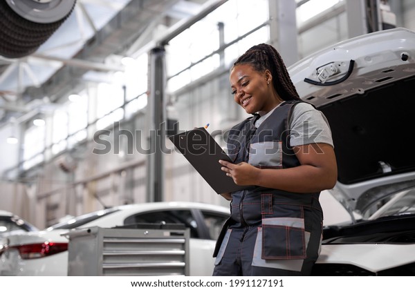 black female Auto Mechanic Making\
Car Checkup, in Uniform Writing and holding Clipboard of Service\
Order Indoors. Repair service. Woman Car Repairing\
Concept