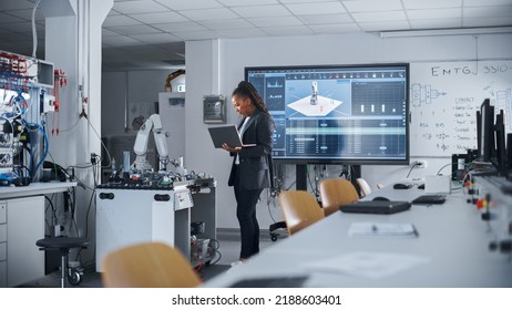 Black Female Automation Engineer Holding Laptop And Moving Bionic Arm With Written Code At Futuristic Factory. Modern Equipment And New Era In Computer Science And Robotics Industry Concept.