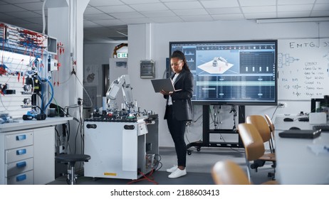 Black Female Automation Engineer Holding Laptop And Moving Bionic Arm With Written Code At Futuristic Factory. Modern Equipment And New Era In Computer Science And Robotics Industry Concept.