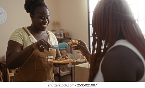 Black female baker smiling while serving a pastry to a customer inside a cozy bakery shop with a warm interior. - Powered by Shutterstock