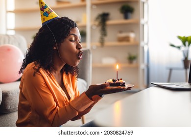 Black Female Blowing Candle On Birthday Cake Having Online B-Day Party, Wearing Festive Hat At Home. Side View Shot. Modern Distance Holiday Celebration Concept