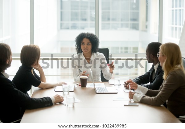 Black female boss leading corporate multiracial\
team meeting talking to diverse businesspeople, african american\
woman executive discussing project plan at group multi-ethnic\
briefing in boardroom