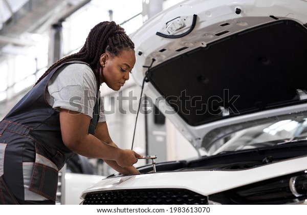 Black Female car mechanic\
holding wrench checking up on the car engine, for repair and\
checkup, wearing overall, repairing auto hood. side view portrait.\
copy space