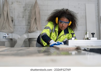 Black female carpenter wearing ear protection glasses and gloves safety using circular saw cutting piece of wood at furniture factory - Powered by Shutterstock