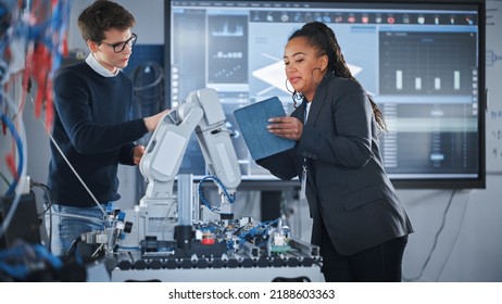 Black Female Chief Engineer Holding Tablet And Talking With Male Student While Robot Arm Moving Under Their Supervision During The Lesson. Computer Science Education And Learning Concept.