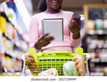 Black Female Consumer With Shopping Trolley Full Of Products Checking Grocery List, Choosing Products At Supermarket. Unrecognizable African American Lady Buying Food At Mall, Low Angle View