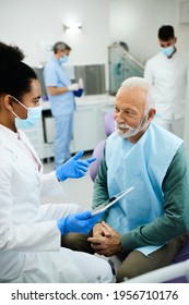 Black Female Dentist Talking To Senior Patient After Dental Exam At Dentist's Office. Focus Is On Man.