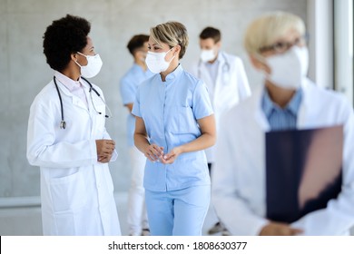 Black Female Doctor And Nurse With Protective Face Masks Communicating In A Lobby At Medical Clinic. 