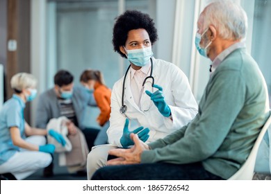 Black female doctor and senior man wearing protective face masks while communicating in a waiting room at hospital. 