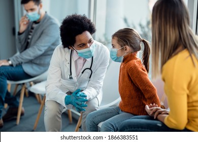 Black Female Doctor Talking To A Small Girl Who Is Sitting With Her Mother At Hospital Waiting Room And Wearing Protective Face Mask Due To Coronavirus Pandemic.