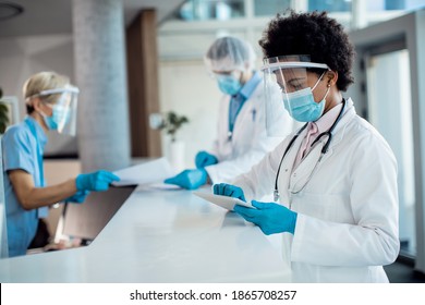 Black Female Doctor Using Digital Tablet While Standing At Reception Desk At Medical Clinic And Wearing Face Shield And Mask Due To COVID-19 Pandemic.