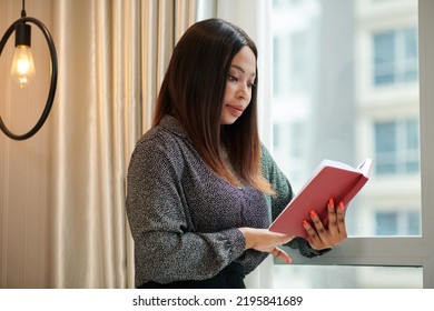 Black Female Entrepreneur Standing At Window And Reading Book On Self Improvement