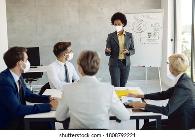 Black female entrepreneur wearing protective face mask while presenting to her coworkers new business ideas on whiteboard in the office.  - Powered by Shutterstock