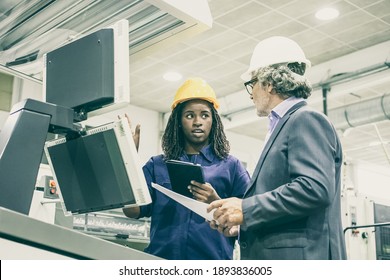Black Female Factory Worker And Her Male Boss Standing At Industrial Machine And Talking. Low Angle. Production Process Or Machinery Concept