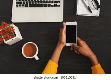 Black female hands holding smartphone with blank screen for advertisement. Top view of african-american hands, laptop keyboard, coffee, mobile on dark wooden table background, copy space