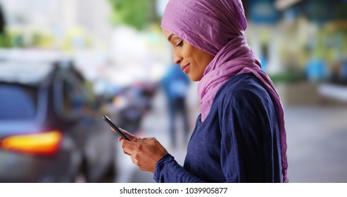 Black female in her 20s wearing hijab texting on cellphone on urban street - Powered by Shutterstock