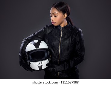 Black Female Motorcycle Biker Or Race Car Driver Or Stuntwoman Wearing Leather Racing Suit And Holding A Protective Helmet.  She Is Standing Confidently In A Studio