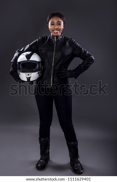Black female motorcycle\
biker or race car driver or stuntwoman wearing leather racing suit\
and holding a protective helmet.  She is standing confidently in a\
studio