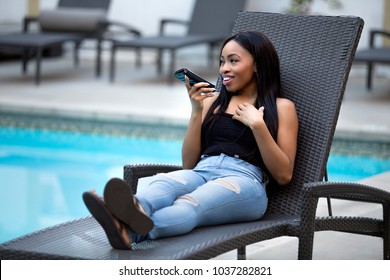 Black Female On A Speaker Phone Call In A Hotel Resort.  She Is Working While On Vacation Or Dictating Reminders On A Voice Assistant On Her Cellphone. The Woman Is Sitting By The Hotel Pool.