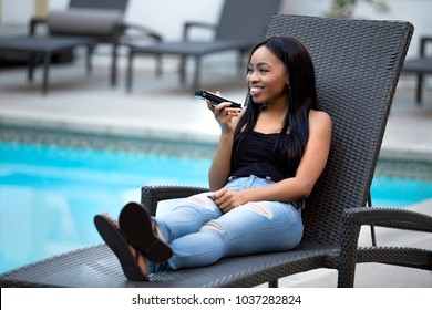 Black Female On A Speaker Phone Call In A Hotel Resort.  She Is Working While On Vacation Or Dictating Reminders On A Voice Assistant On Her Cellphone. The Woman Is Sitting By The Hotel Pool.