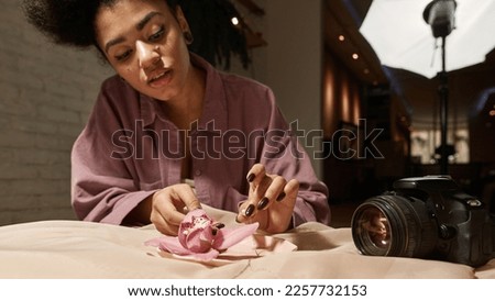 Black female photographer preparing chainlet and pink flower for taking photo on digital camera at home studio. Young girl creating content for photostocks, commerce, social networks and advertising