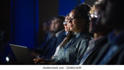 Black Female Sitting in Dark Crowded Auditorium at an International Business Conference. Multiethnic African Woman Using Laptop Computer. Delegate Watching Presentation About New Financial Solutions. - Powered by Shutterstock