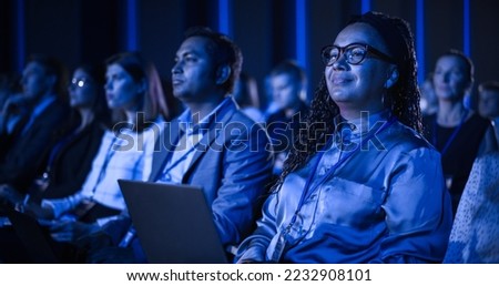 Black Female Sitting in Dark Crowded Auditorium at an International Business Conference. Multiethnic African Woman Using Laptop Computer. Delegate Watching Presentation About New Financial Solutions.