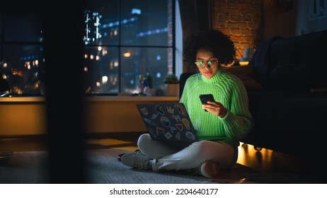 Black Female IT Specialist Coding on her Laptop in the Evening and Checking Notifications on Smartphone. Diverse Woman has Deadline Late at Night. Millennial People Working Concept. - Powered by Shutterstock